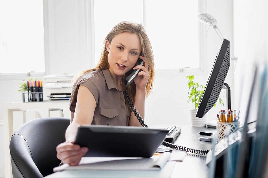 En kvinde med kontaktlinser sidder ved sit skrivebord, kigger på en tablet mens hun taler i telefon.